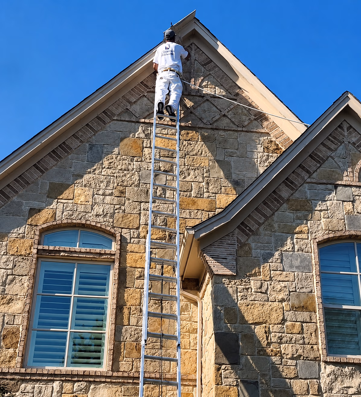 Man in white outfit on a tall ladder working near the roof peak of a stone house under blue sky.