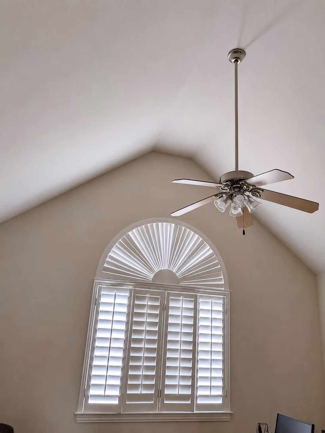 Ceiling fan with wooden blades and lights above a window with white plantation shutters and a half-round fanlight.
